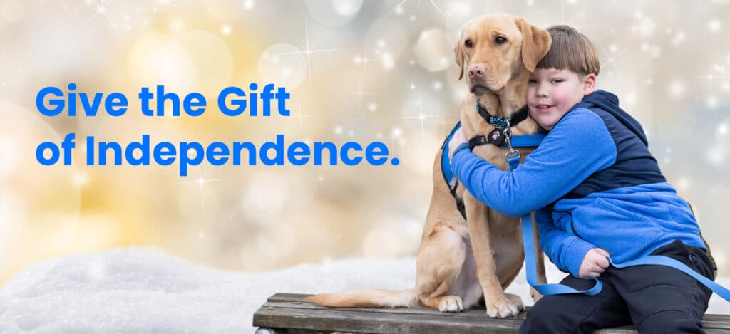 A young boy in a blue jacket hugging a tan service dog wearing a blue harness and leash, seated on a wooden bench with snow in the foreground and a sparkling bokeh background with large blue text reading “Give the Gift of Independence.”