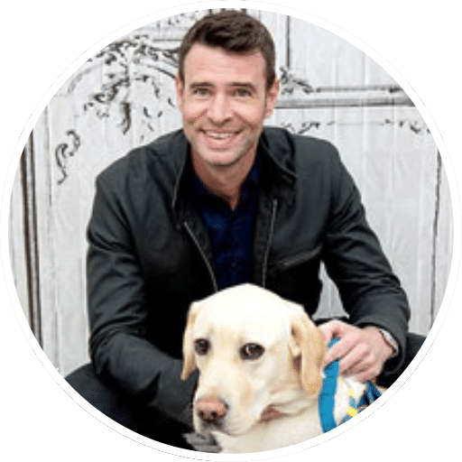 Smiling man in a dark jacket kneeling behind a yellow Labrador wearing a blue service harness in front of a white decorative wooden background.