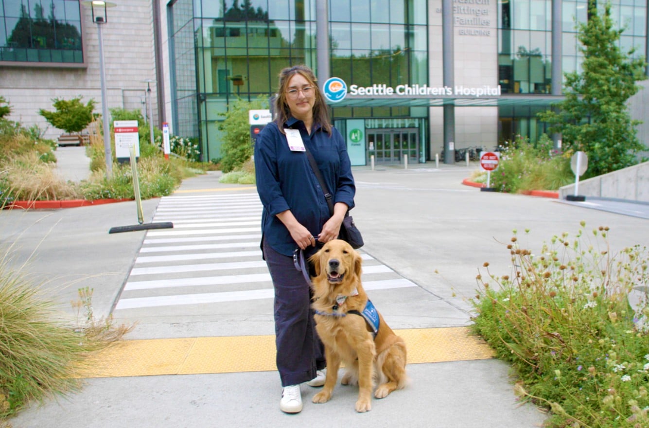 Woman at Seattle Children's Hospital crosswalk holding leash of a seated golden retriever service dog.