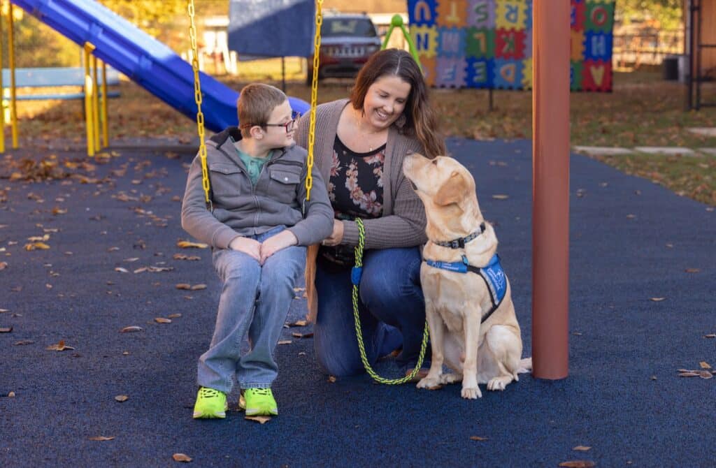 Smiling woman kneeling beside a boy on a swing as a yellow lab in a blue service vest sits nearby