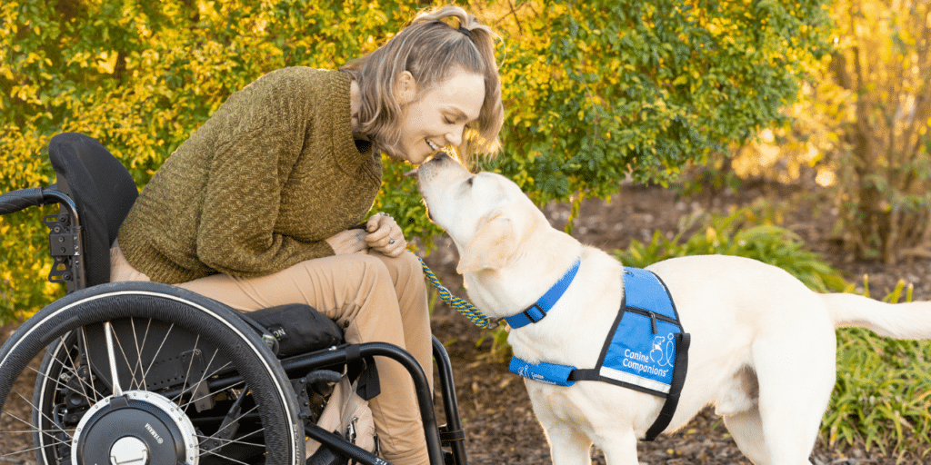 A smiling woman in a wheelchair leans forward as a yellow Labrador wearing a blue Canine Companions service vest nuzzles her face in a sunlit garden.