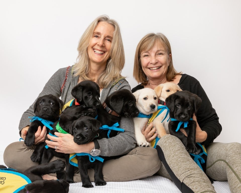 Two smiling middle-aged women sitting against a white background, each holding a cluster of black and yellow Labrador puppies wearing bright blue and yellow bandanas.