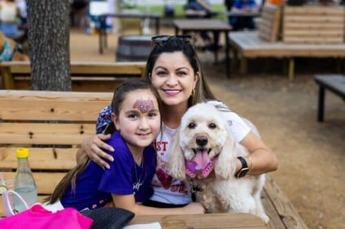 Smiling woman embraces a girl with face paint and their cream-colored dog wearing a pink bandana.