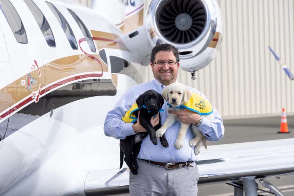 Man smiling in front of a private jet holding a black and a yellow Labrador puppy wearing yellow training vests