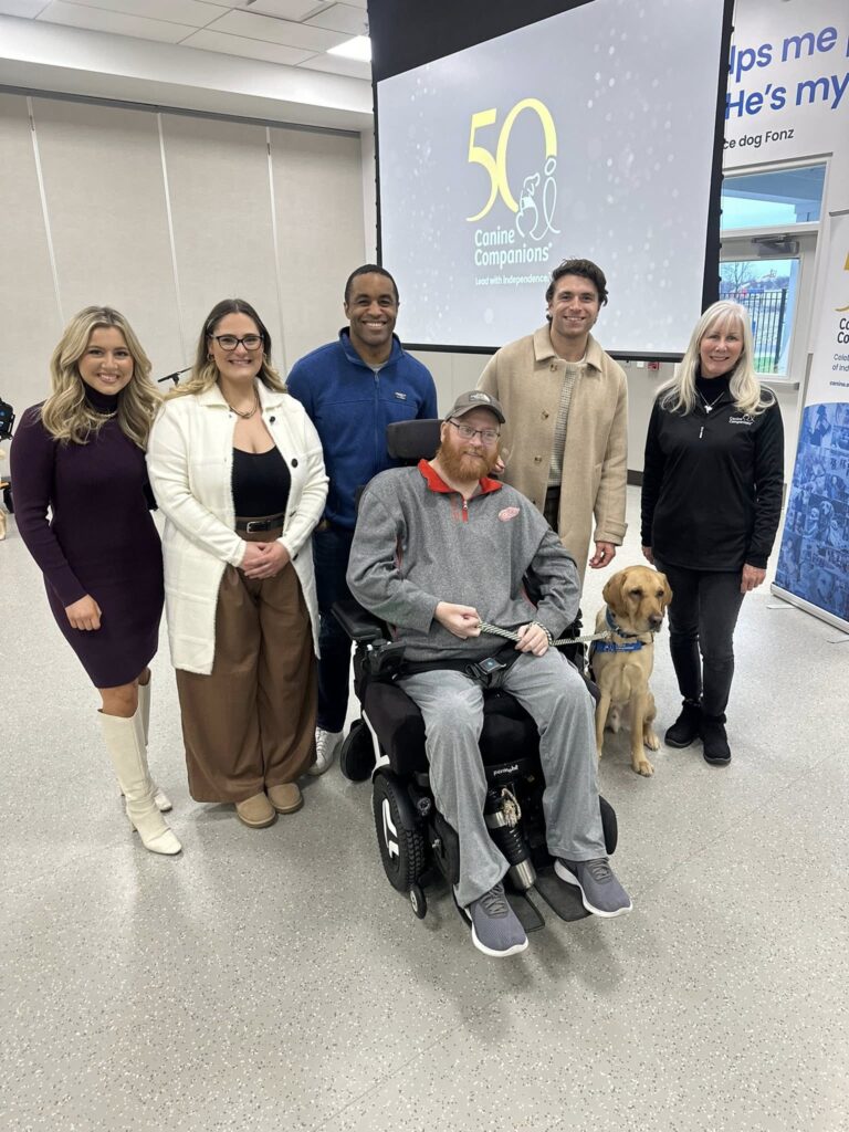 Group of six adults around a man in a power wheelchair holding a yellow service dog, with a 50 Canine Companions logo projected behind.
