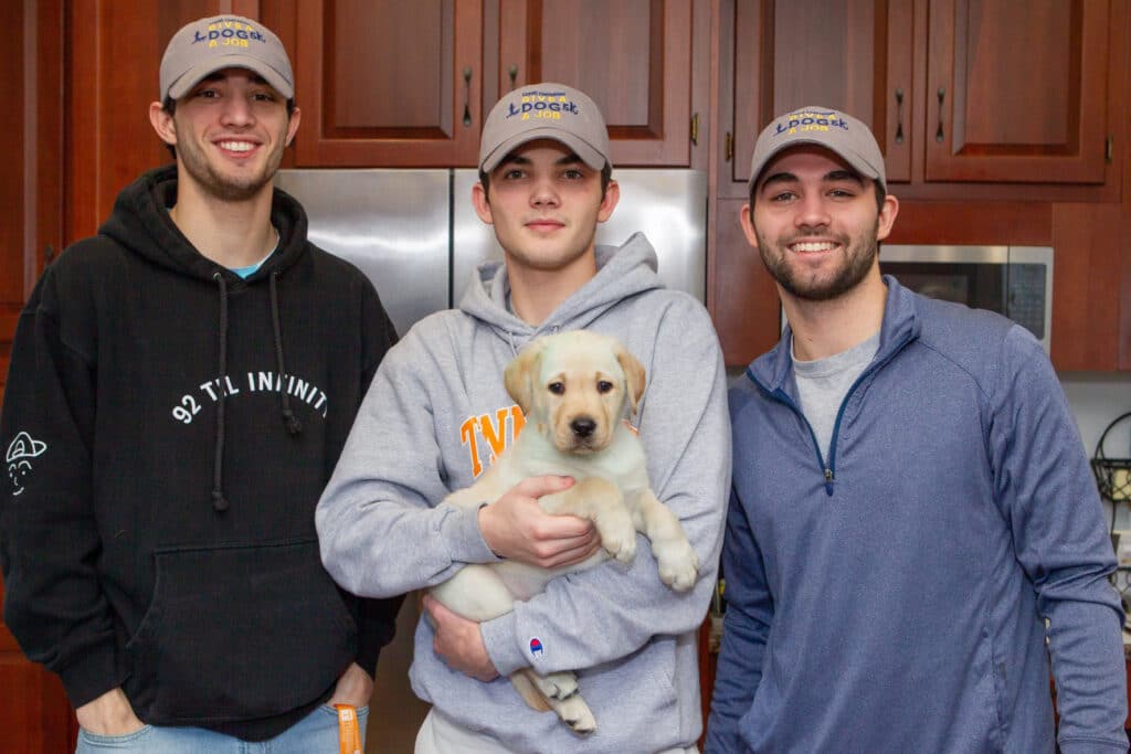 Three smiling young men wearing matching caps stand in a kitchen, the center person holding a light-colored Labrador puppy