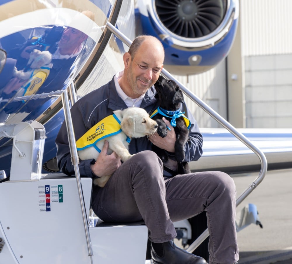 Man sitting on jet stairs holding a yellow and a black puppy in Canine Companions vests, black puppy nuzzling his face.