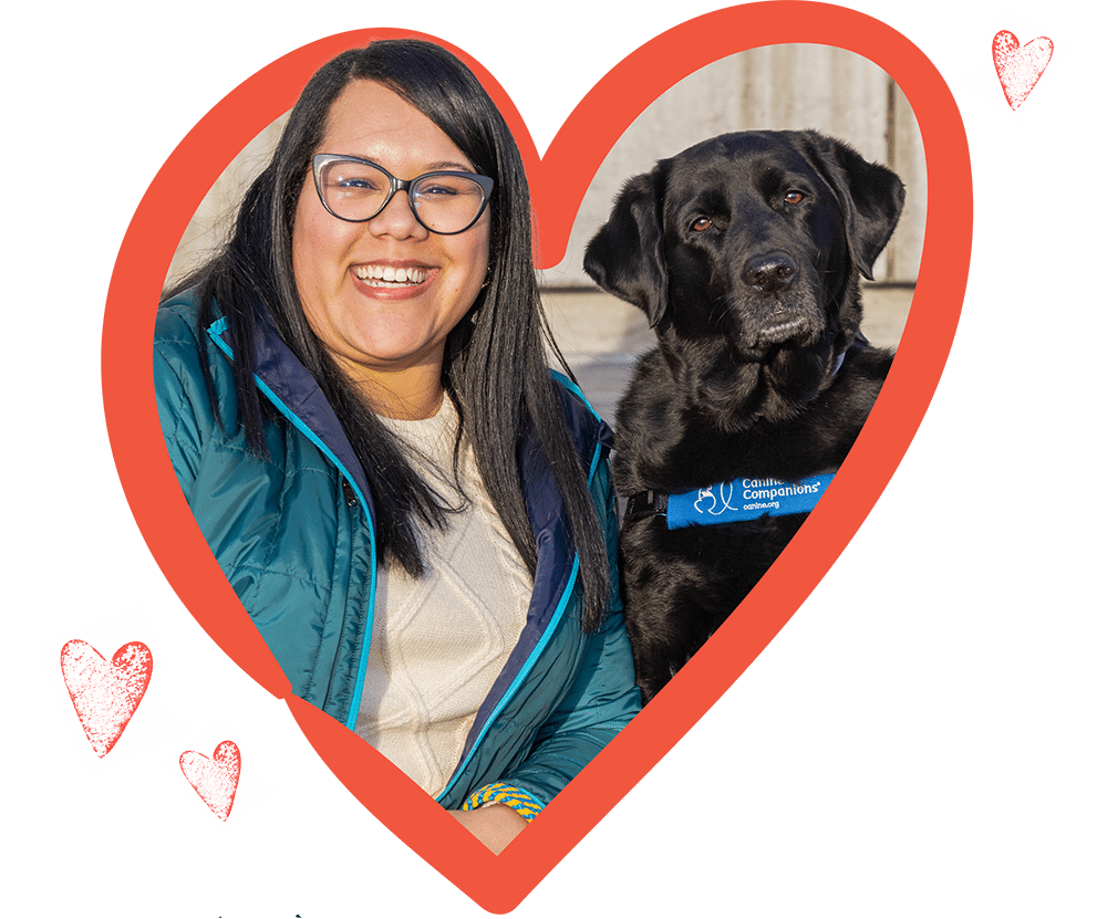 woman smiling with glasses beside a black Labrador wearing a blue service vest, both framed by a red heart graphic