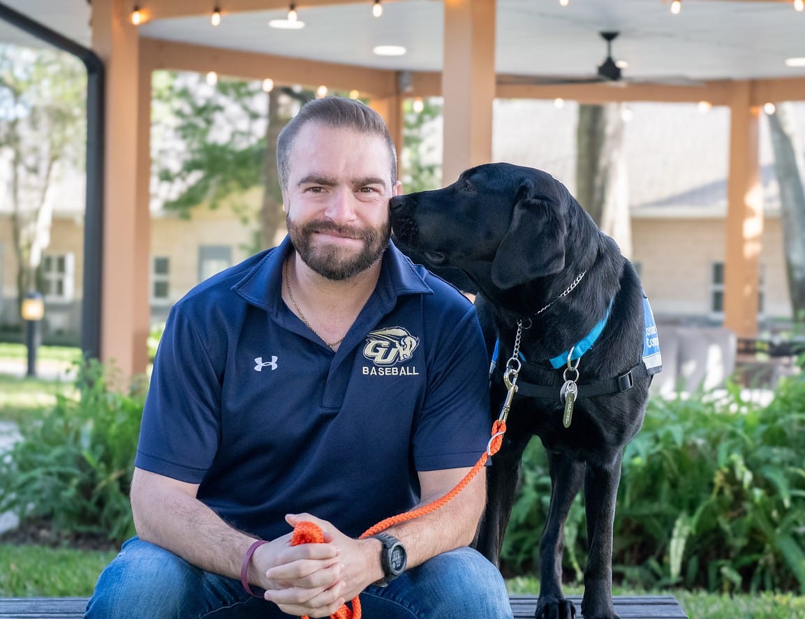 Bearded man in navy baseball polo smiles as a black service dog wearing a blue vest nuzzles his face, orange leash visible.