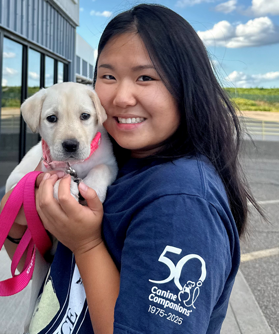 Woman smiling, holding a white puppy with a pink collar and leash, wearing a navy shirt with a '50' Canine Companions logo.