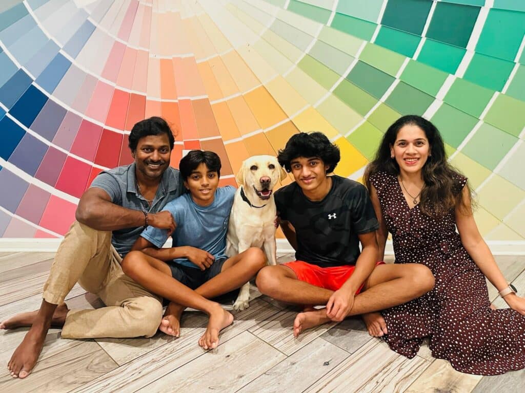 Smiling family of four with a yellow Labrador seated barefoot on a wood floor in front of a colorful paint-swatch wall.