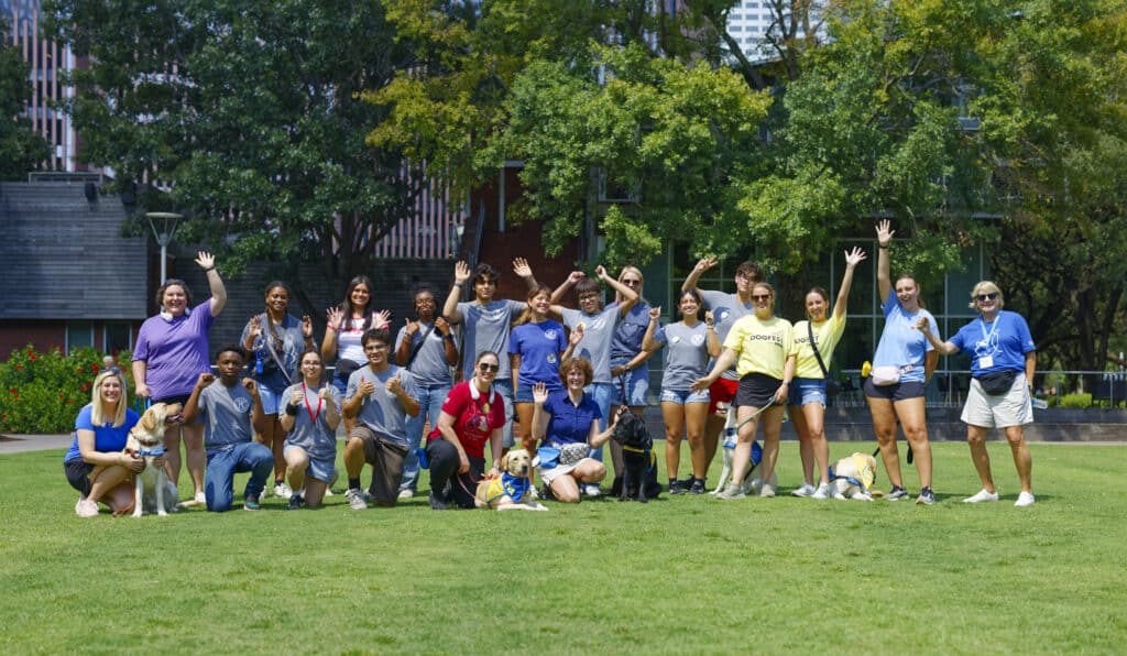 Group of people on a grassy lawn waving and posing with several service dogs in vests, trees and buildings behind.