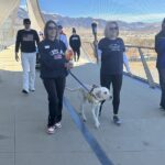 Two women walk a guide dog across a mesh-sided pedestrian bridge with mountains in the background, one holding a lit torch.
