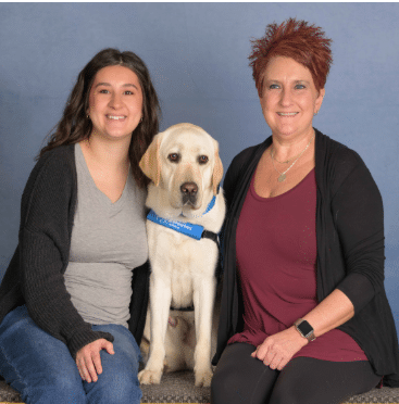 Two smiling women sit on either side of a yellow Labrador wearing a blue service harness against a plain studio backdrop.