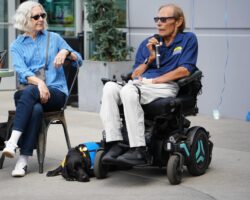 Man in a wheelchair speaking into a microphone with a black service dog in a blue vest at his feet, woman seated nearby.