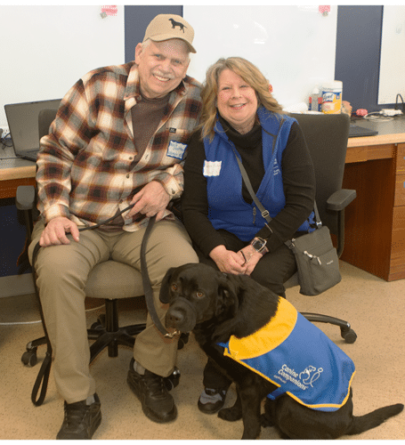 Smiling man in plaid and woman in blue vest seated with a black lab wearing a blue-and-yellow service dog vest.