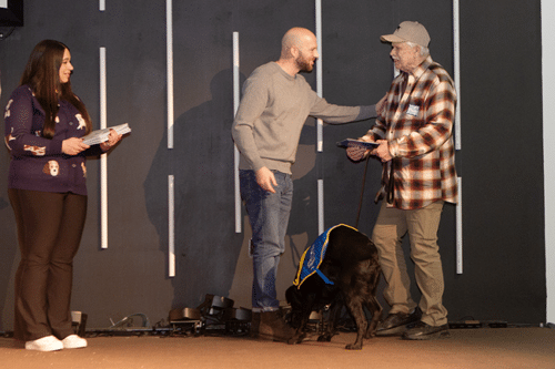 Three people on stage and a black service dog in a blue vest; a man in gray greets a man in plaid and cap.