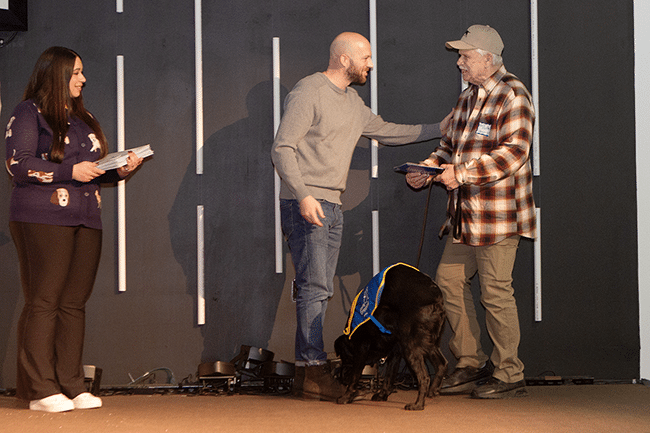 Three people on stage and a black service dog in a blue vest; a man in gray greets a man in plaid and cap.