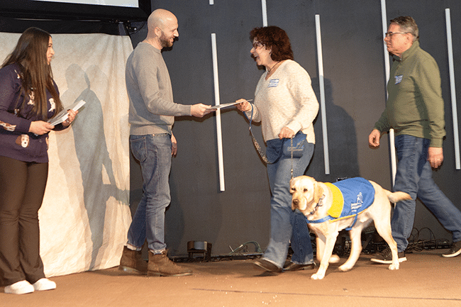 Woman with a yellow Labrador guide dog in a blue service vest accepts a certificate from a man on stage while others look on.