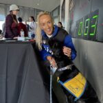 Smiling blonde woman in blue jacket kneeling and petting a black service dog wearing a yellow Canine Companions vest.