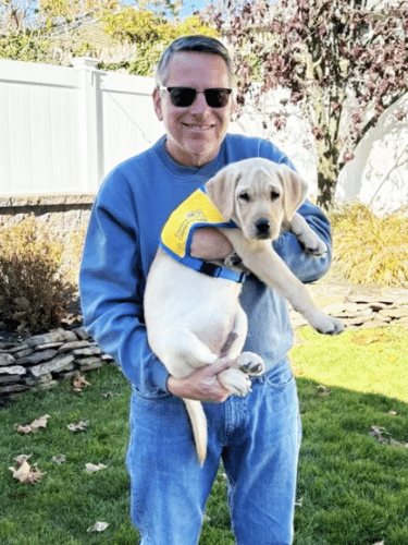 Smiling man in sunglasses and blue sweatshirt holding a yellow Labrador puppy wearing a small yellow vest in a sunny backyard