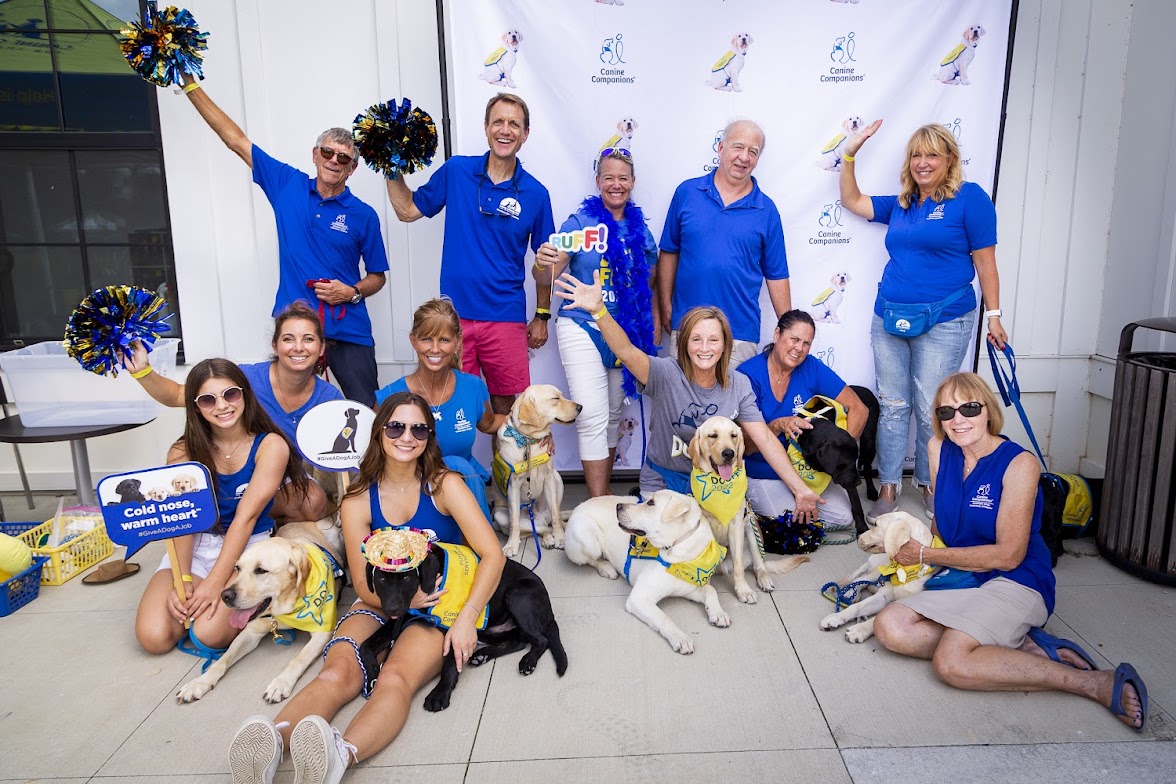 Volunteers in blue with yellow‑vested service dogs, holding pom‑poms and signs before a Canine Companions backdrop.