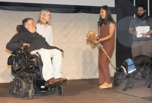Man in power wheelchair smiling at a young woman holding a stuffed bear and a football while a service dog sniffs nearby.