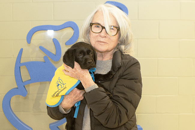 Older woman with white hair and glasses holding a black Labrador puppy wearing a yellow Canine Companions vest in front of a tiled wall with a blue dog silhouette.