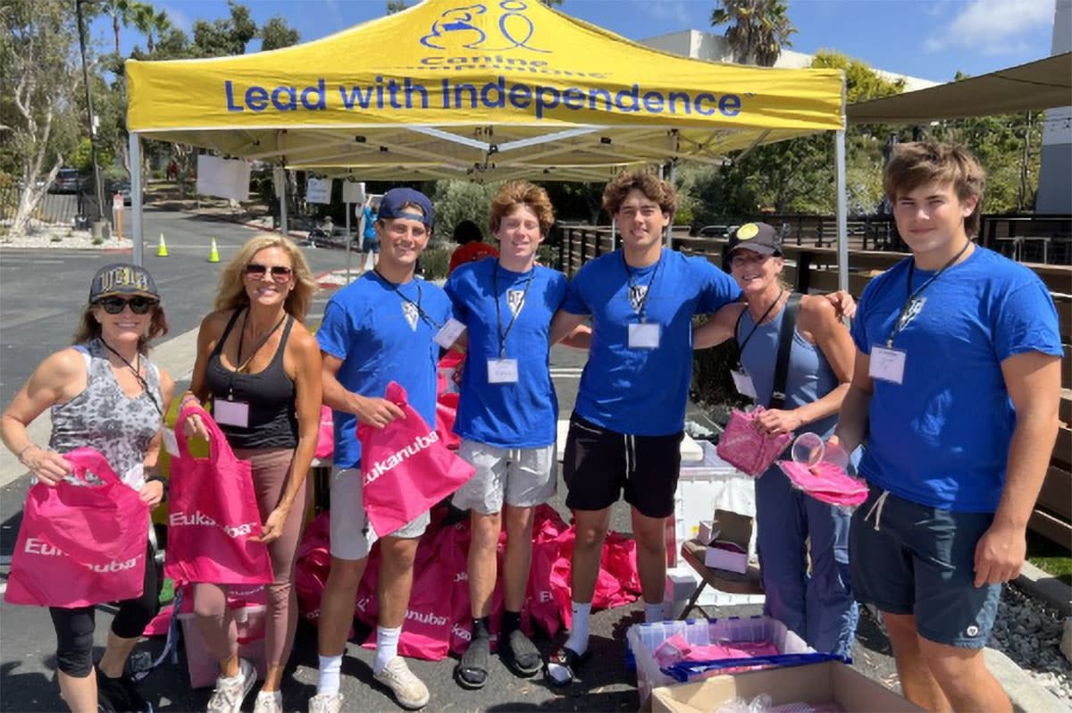 Group of volunteers in blue shirts posing under a yellow canopy that reads Lead with independence while holding pink tote bags.