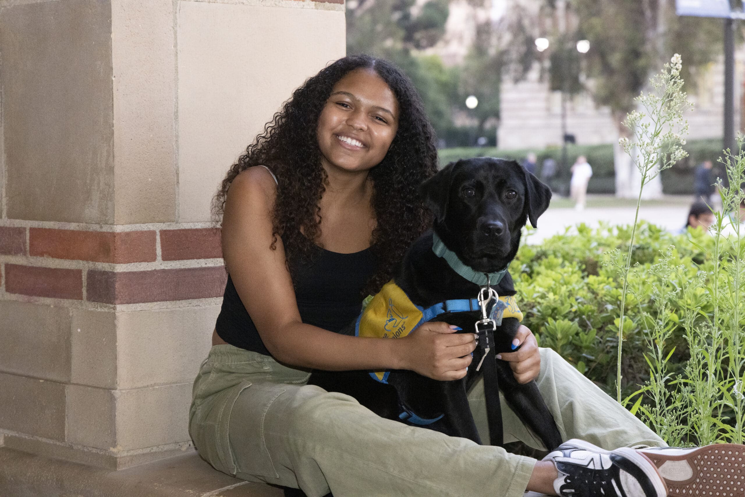 Smiling young woman with curly hair sitting by a brick column, holding a black service dog wearing a yellow vest and teal harness
