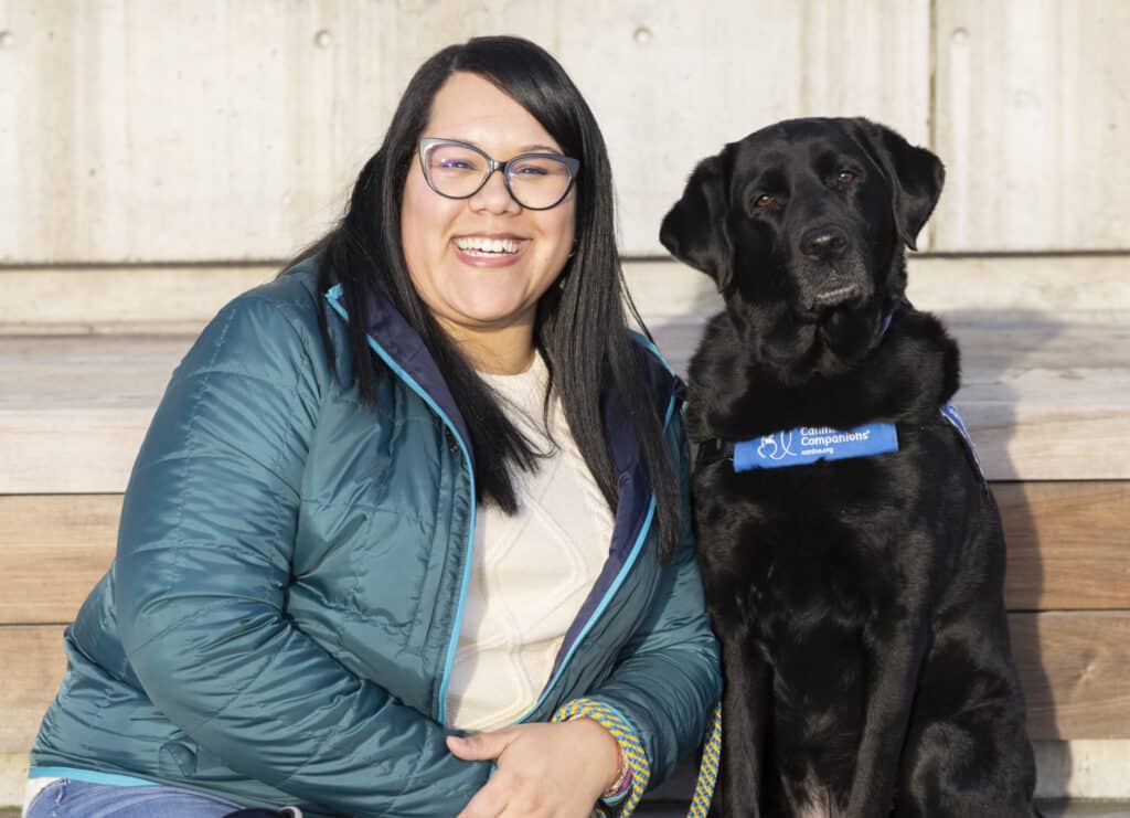 Smiling woman in glasses and teal jacket sits on wooden steps next to a black service Labrador wearing a blue "Canine Companions" vest.