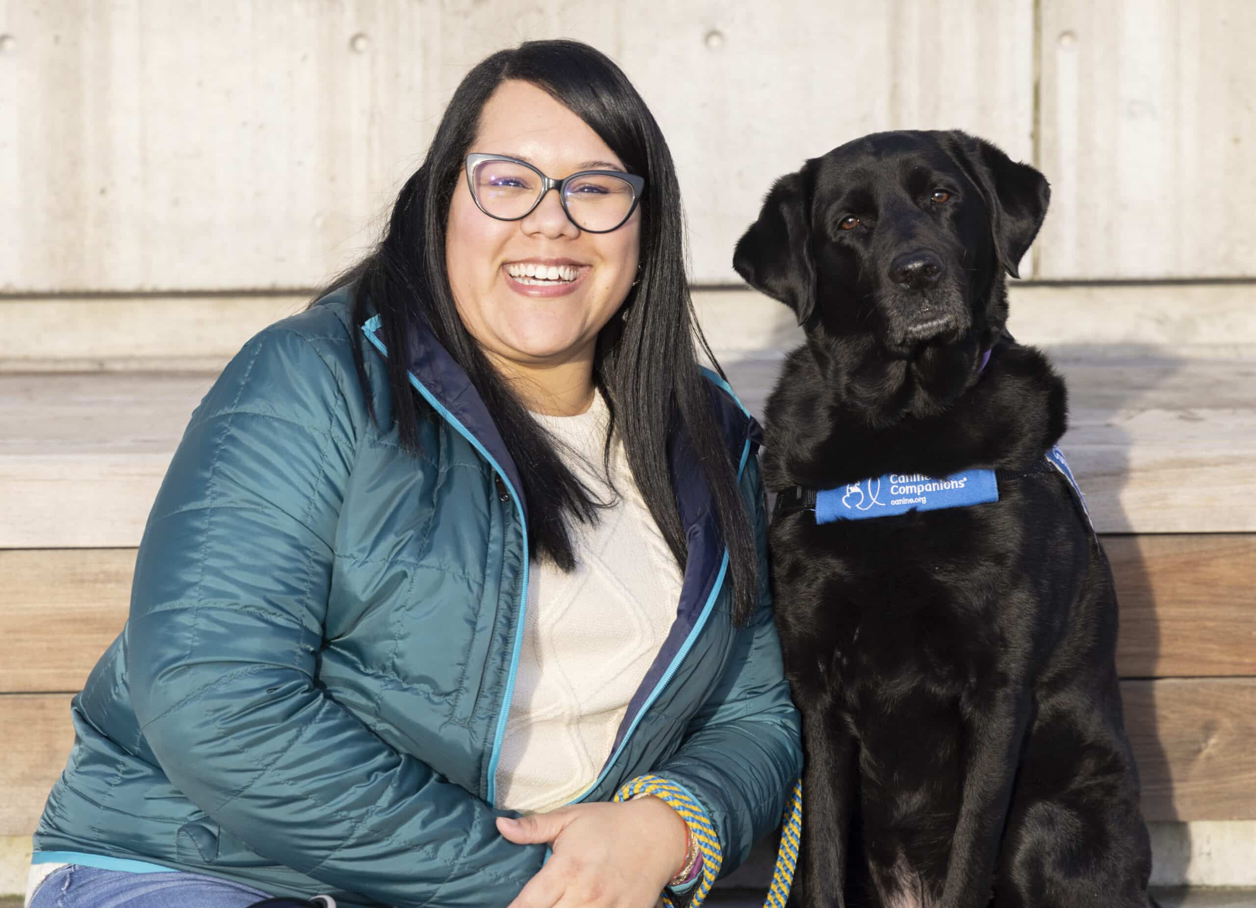 Smiling woman in glasses and teal jacket sits on wooden steps next to a black service Labrador wearing a blue "Canine Companions" vest.