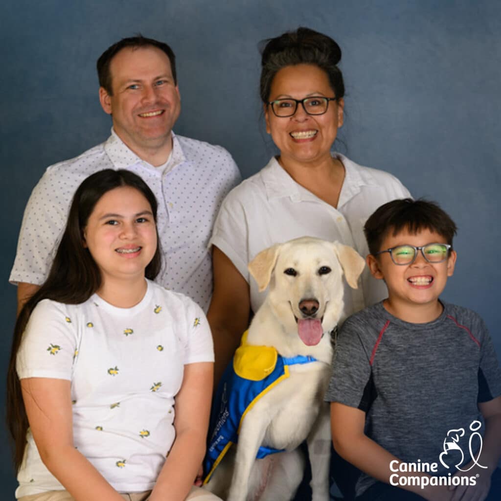 Smiling family portrait with two adults and two children posing with a tan assistance dog wearing a blue-and-yellow service vest and tongue out