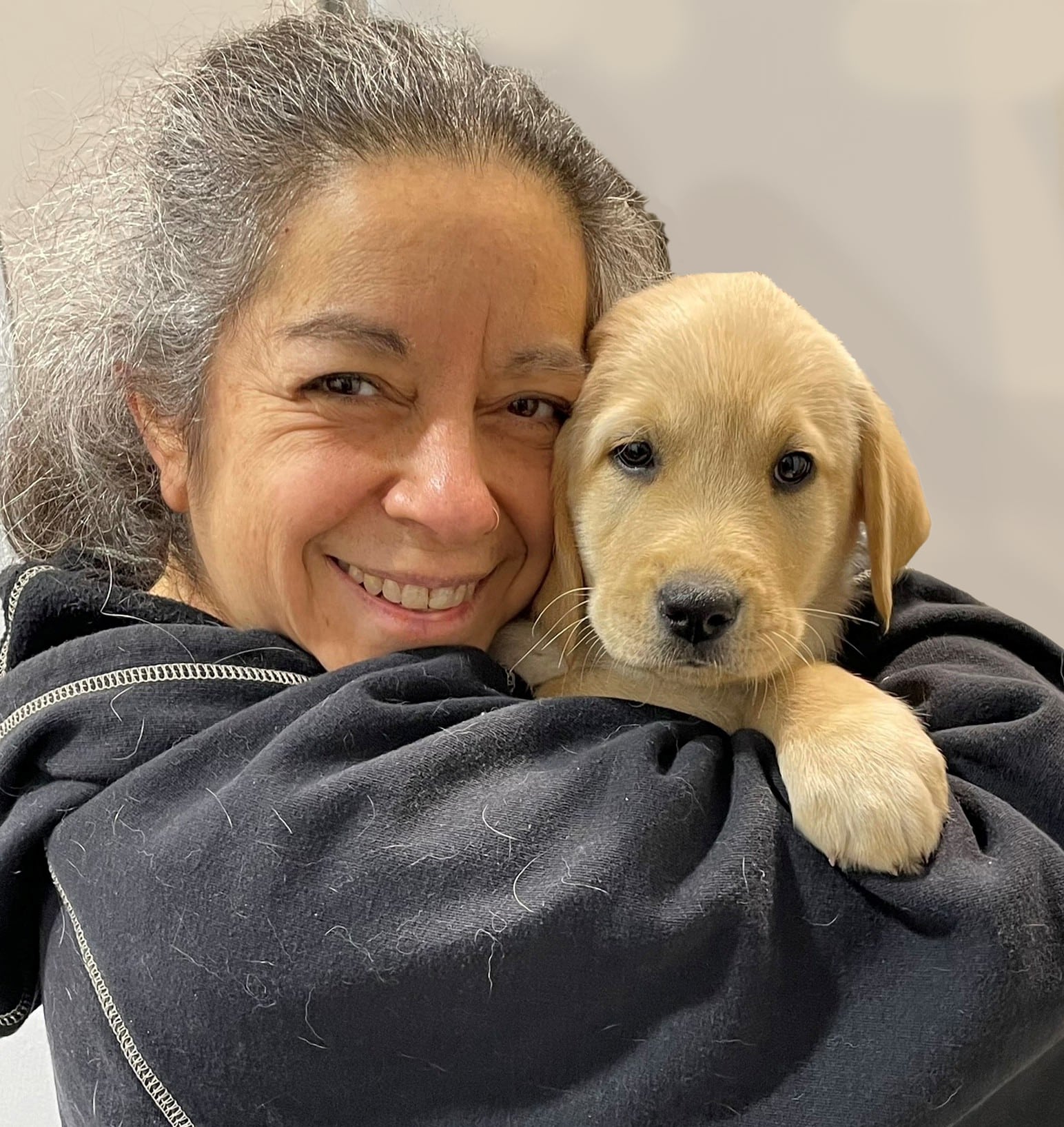 Smiling woman with gray hair hugging a golden Labrador puppy close to her face, both looking at the camera, woman wearing a dark sweatshirt with pet hair.