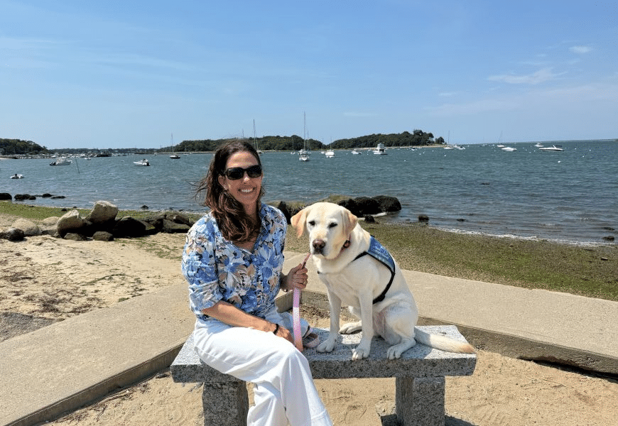 Woman in sunglasses smiling on a stone bench by the water holding a yellow Labrador wearing a blue service vest and pink leash