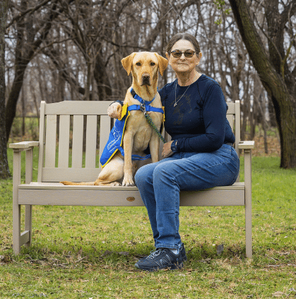 Woman wearing glasses sits on a park bench with a yellow Labrador in a blue service-dog vest beside her, leafless trees in the background.