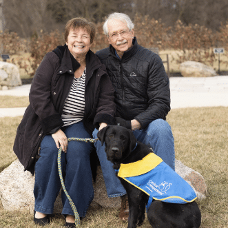 Smiling older couple sitting on a rock outdoors with a black Labrador wearing a blue-and-yellow service dog vest in front of them