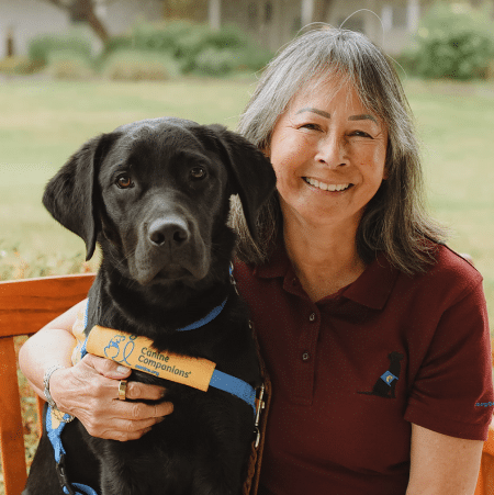 Smiling older woman with gray hair sitting outdoors, holding a black Labrador wearing a yellow "Canine Companions" service vest.
