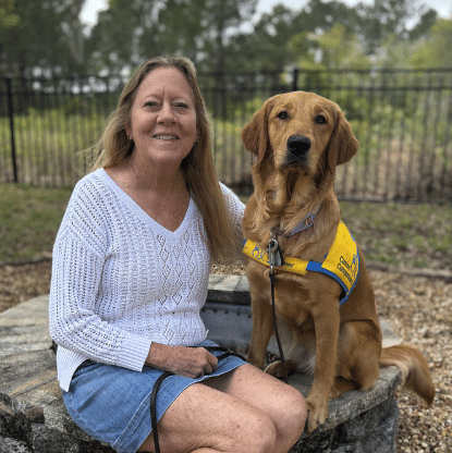 Woman sitting on a low stone wall outdoors beside a golden retriever wearing a yellow service dog vest, both facing the camera