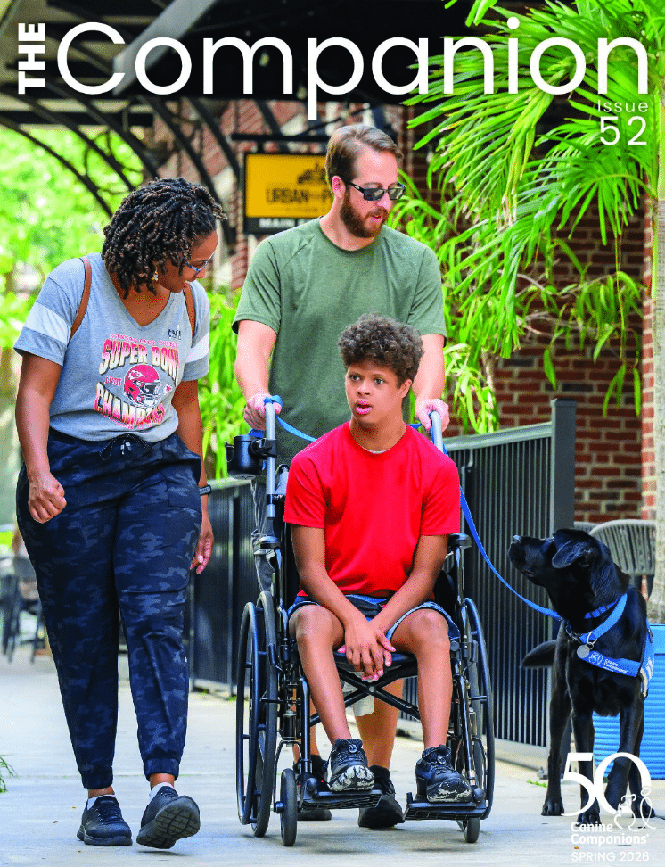 Teen boy in a red shirt in a wheelchair pushed by a man, accompanied by a woman walking and a black service dog on a city sidewalk
