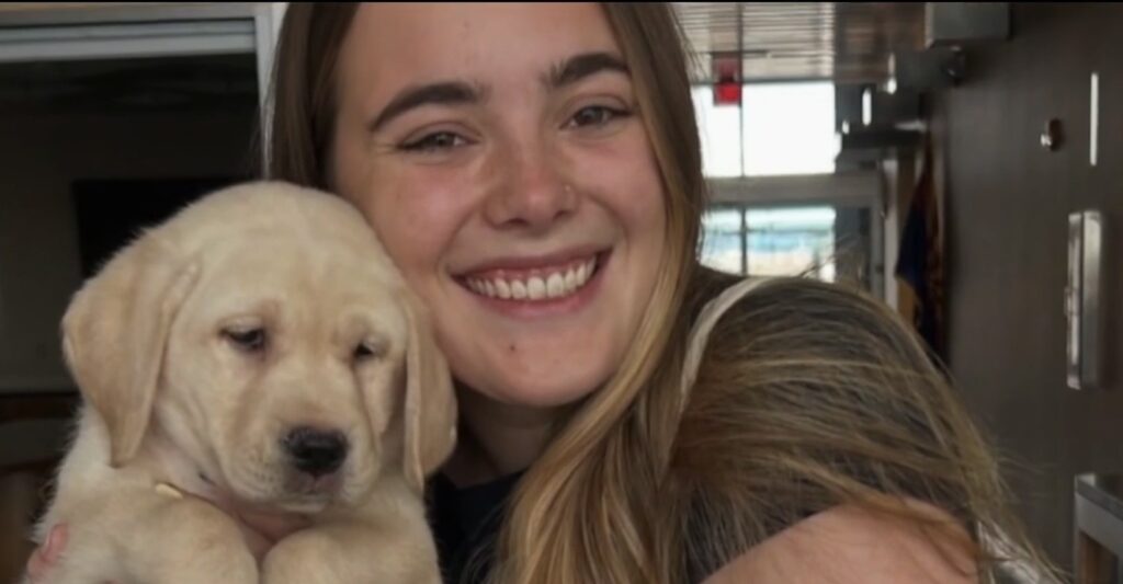 Young woman smiling while hugging a light yellow Labrador puppy close to her face indoors