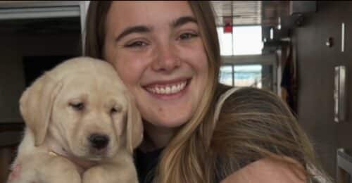 Young woman smiling while hugging a light yellow Labrador puppy close to her face indoors
