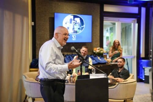 Smiling man in a dress shirt speaking into a microphone at a podium during an indoor event with seated guests.