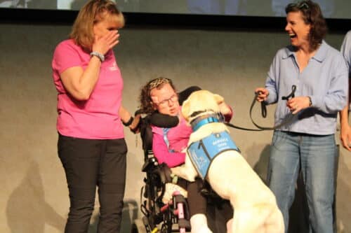 Young woman in a wheelchair wearing pink being greeted enthusiastically by a yellow service dog in a blue vest while two women nearby smile and laugh