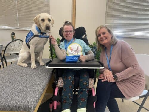 Young woman in a wheelchair smiling with a seated white service Labrador wearing a blue vest and a woman crouching beside them in a classroom-like room
