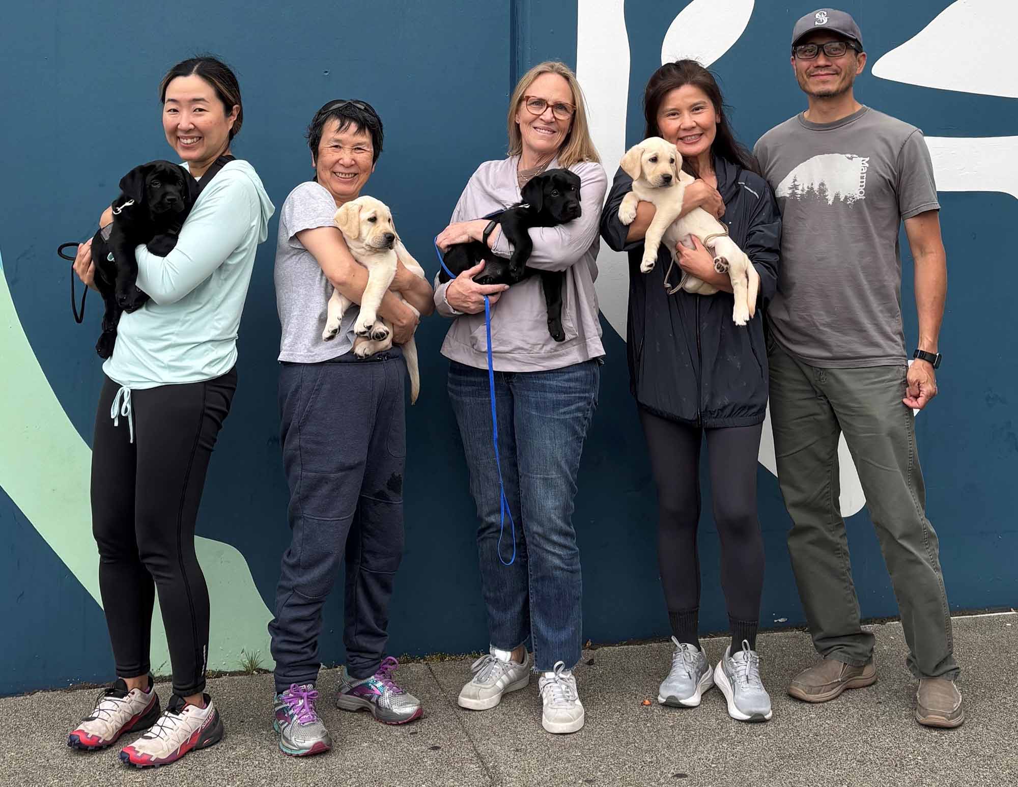 Five people standing in front of a mural, each holding a Labrador puppy (two black, three yellow), smiling.