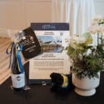 Gift basket with wine, a plant auction sign, white orchids, and a puppy in a yellow vest