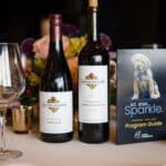 Wine bottles and glasses beside a Canine Companions program guide featuring a puppy in a blue vest