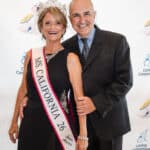 Smiling crowned woman in black gown and sash stands with a man in a suit at a Canine Companions event backdrop