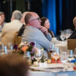 Audience seated at a banquet table listening to a speaker, with floral centerpieces and blue drapes in the background